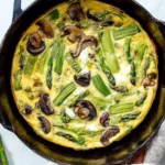 Mushroom and asparagus frittata topped with creamy goat cheese in a skillet